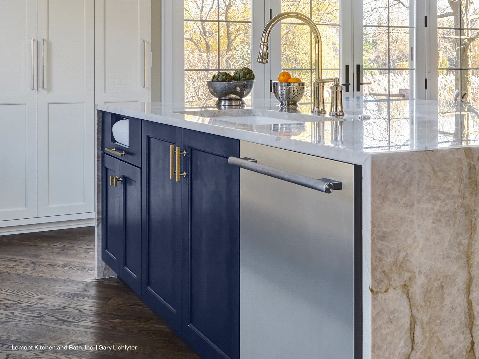 Kitchen island with navy-blue cabinets, waterfall marble countertop, integrated stainless dishwasher panel, chrome faucet and fruit bowl by large windows.