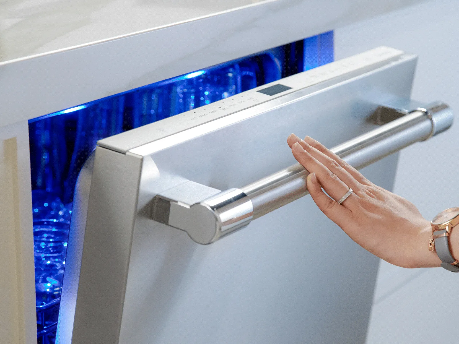 Hand reaching to close a stainless-steel dishwasher door, blue-lit interior with glassware visible.