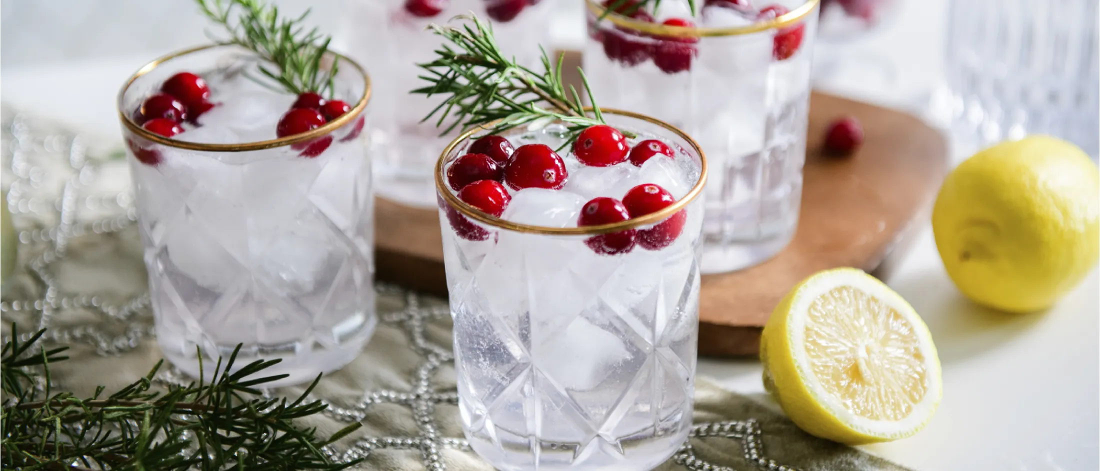 Four crystal glasses with gold rims filled with ice, cranberries, and rosemary sprigs, next to a wooden board and halved lemon