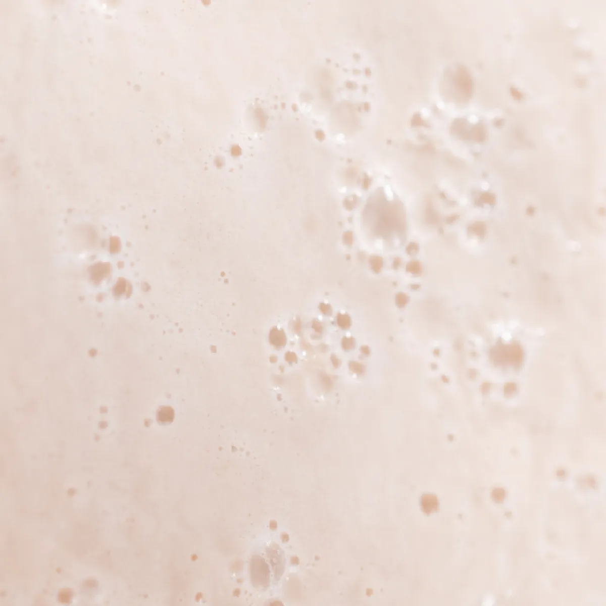 Close-up of a bubbly, creamy beige liquid surface with small clusters of foam and bubbles.