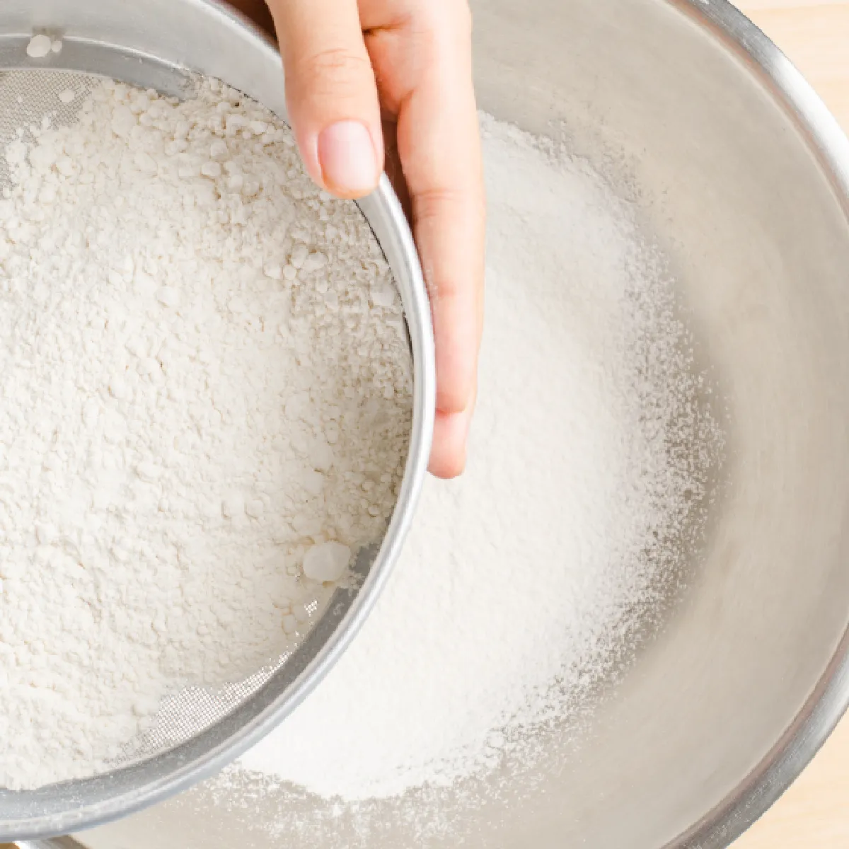 Hand sifting flour through a metal sieve into a large mixing bowl on a light wooden surface.