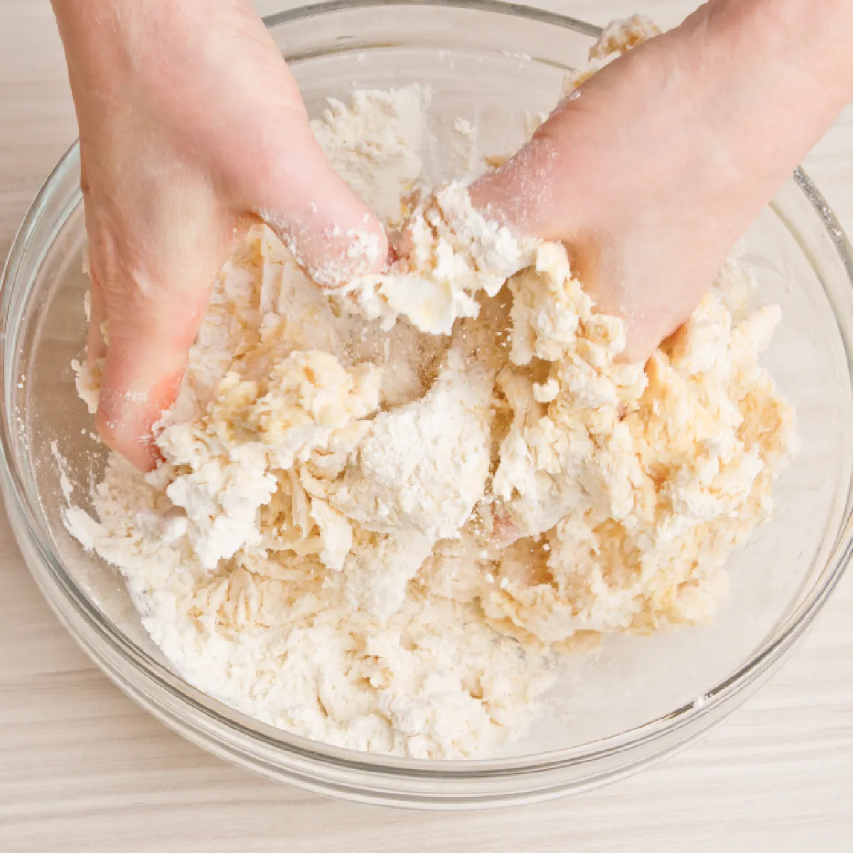 Hands mixing flour and dough in a clear glass bowl on a light wooden surface.