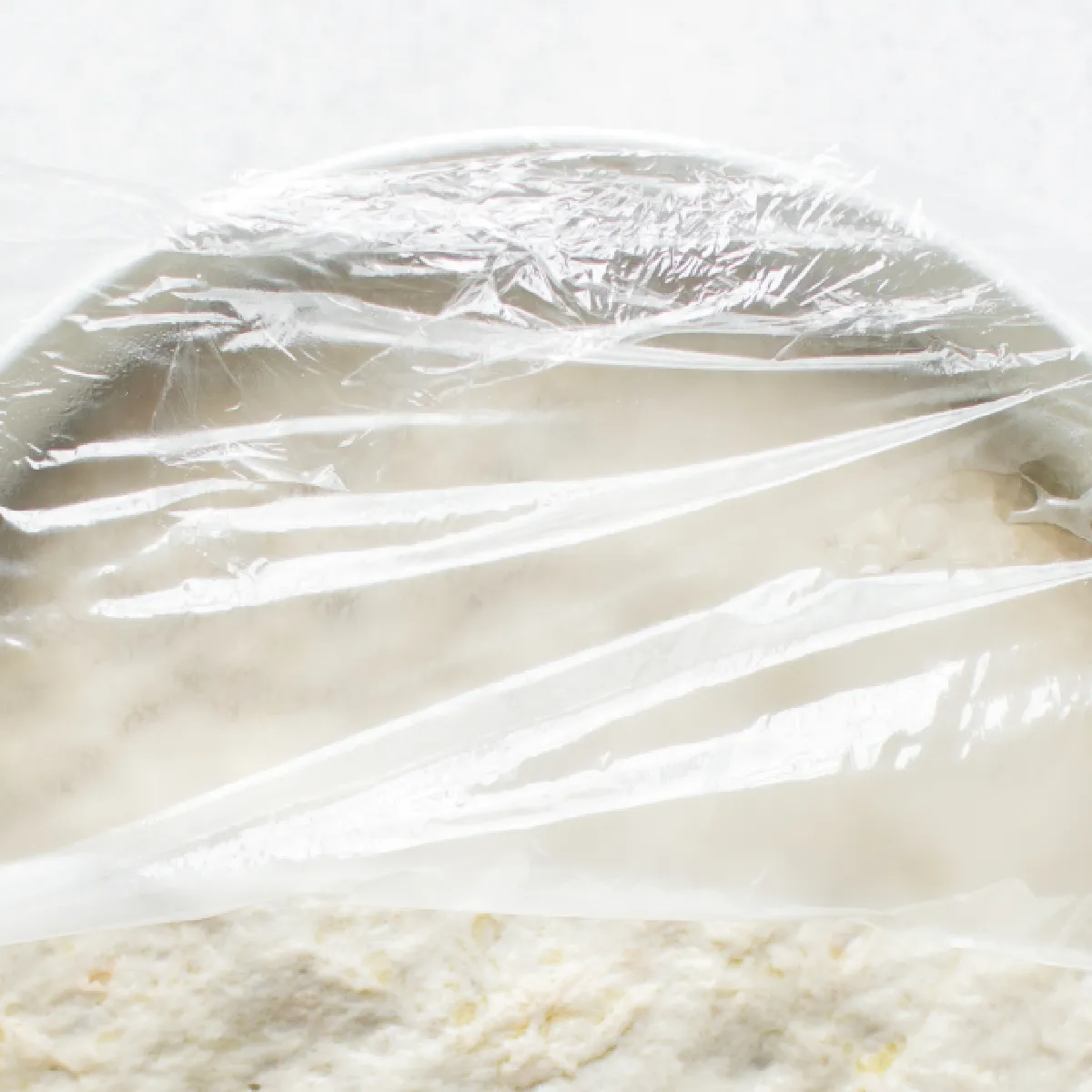 A bowl of dough covered with plastic wrap, shown from above, resting on a white surface.