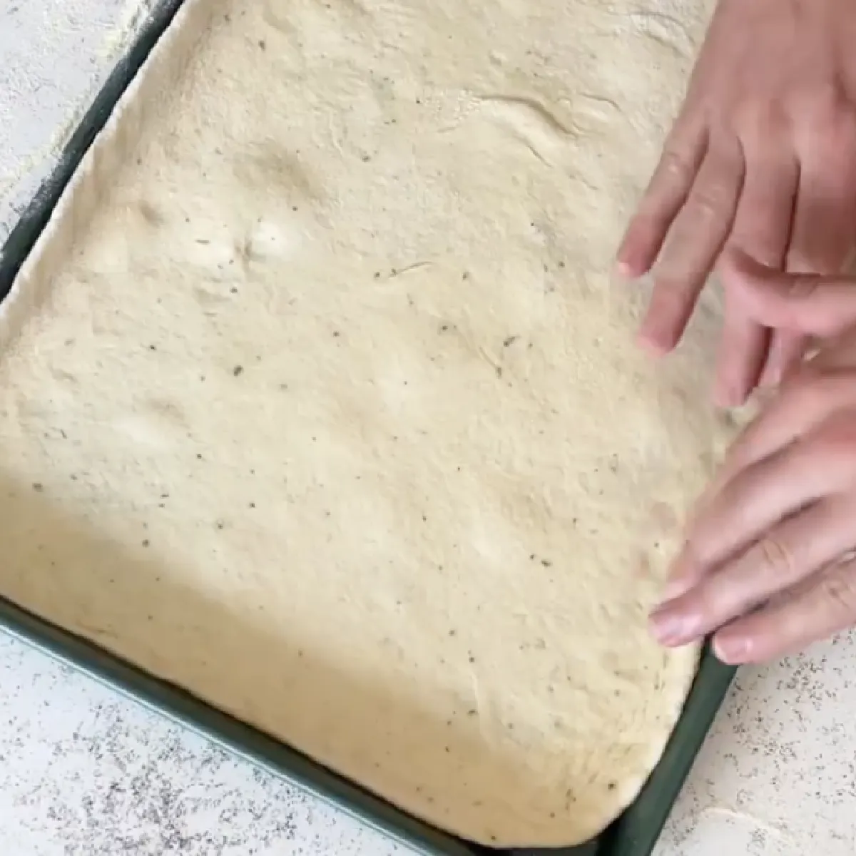 Hands pressing and stretching soft rectangular pizza dough in a green baking tray on a lightly floured surface, preparing homemade pan pizza crust.
