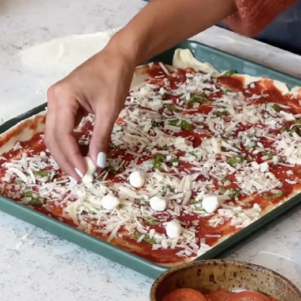 Hand sprinkling shredded mozzarella cheese and herbs over a rectangular sheet pan pizza with tomato sauce in a green baking tray.