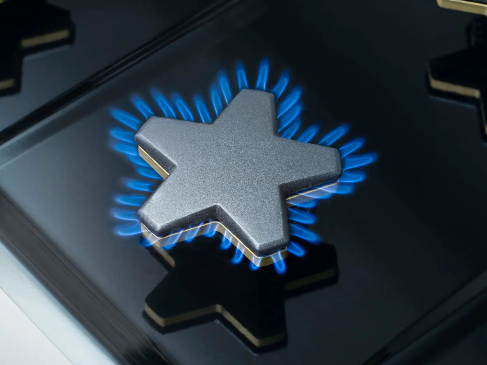 A close-up of a black gas stovetop burner with a unique star-shaped grate. Bright blue flames are visible around the edges of the star, indicating the burner is on. The surface of the stovetop is reflective.