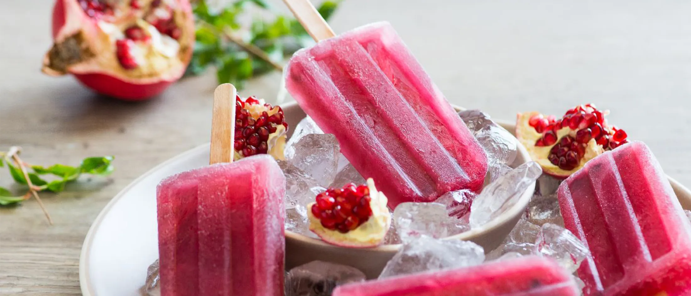 Four pink pomegranate popsicles on ice in a white bowl with pomegranate seeds and green leaves in the background.