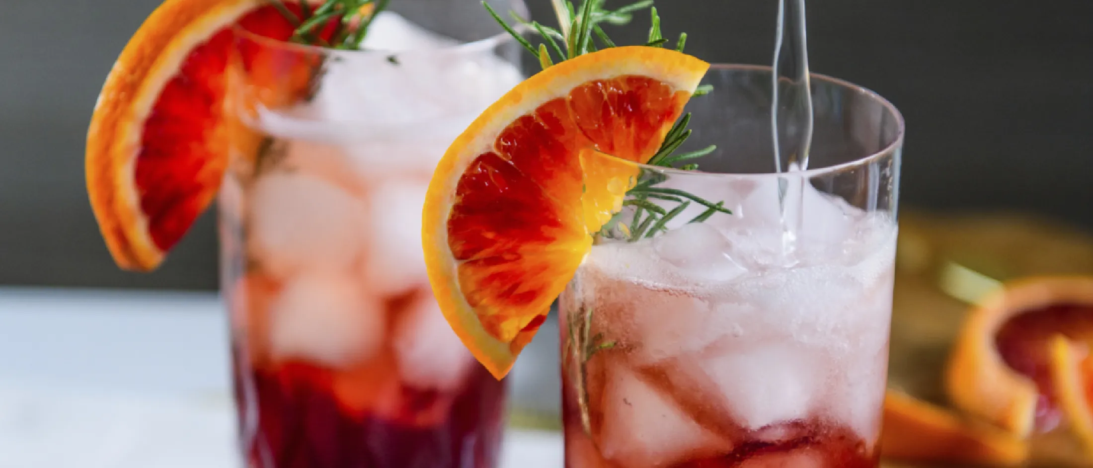 Two iced beverages garnished with blood orange slices and rosemary sprigs, one being poured with a clear liquid.