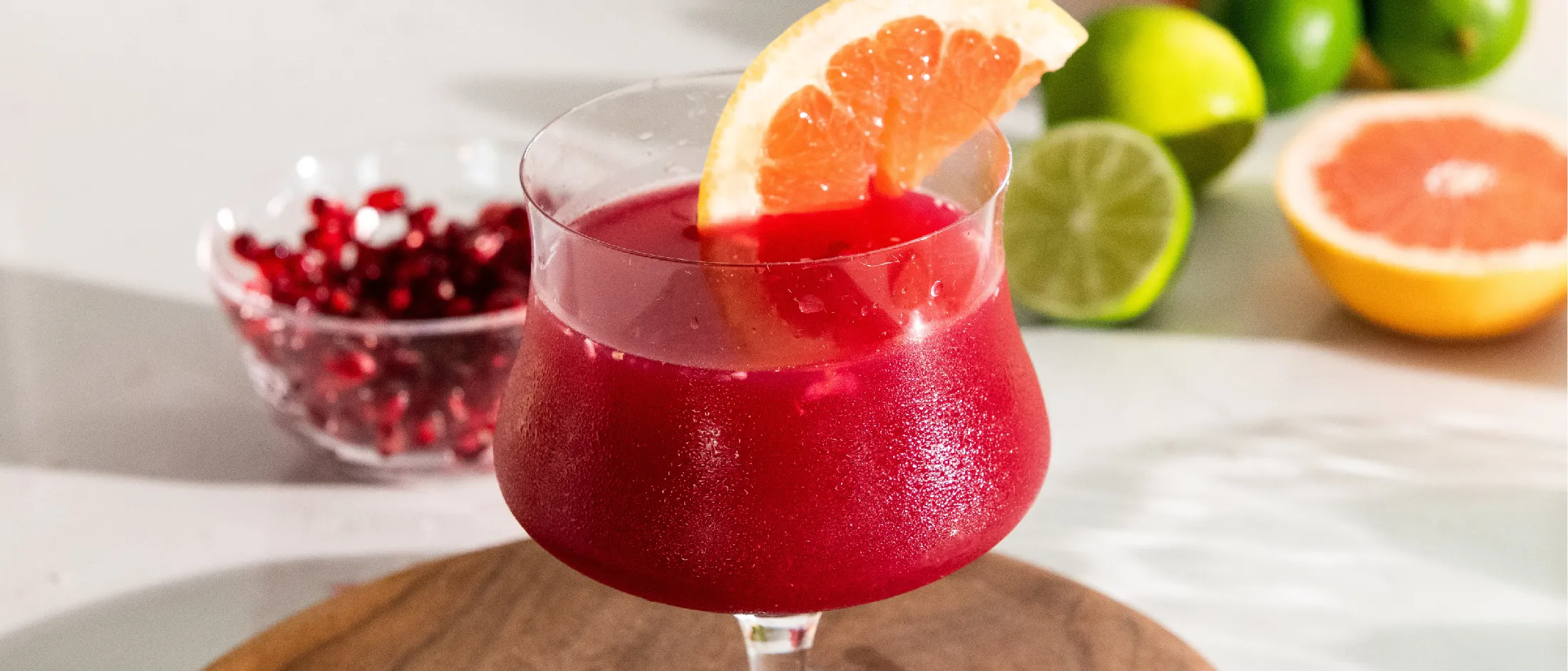 Red pomegranate drink in a glass with a grapefruit slice garnish, with citrus fruits and pomegranate seeds in the background.