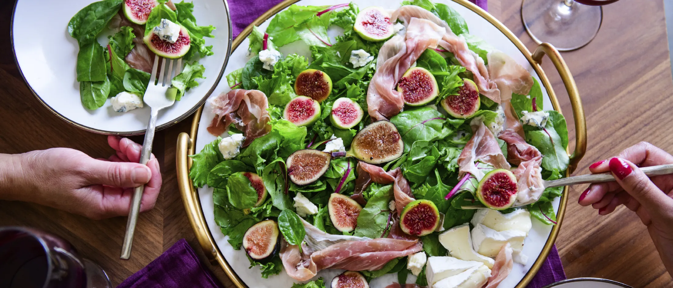 Green salad with figs, prosciutto, blue cheese, and brie, served with red wine on a wooden table with two people eating.