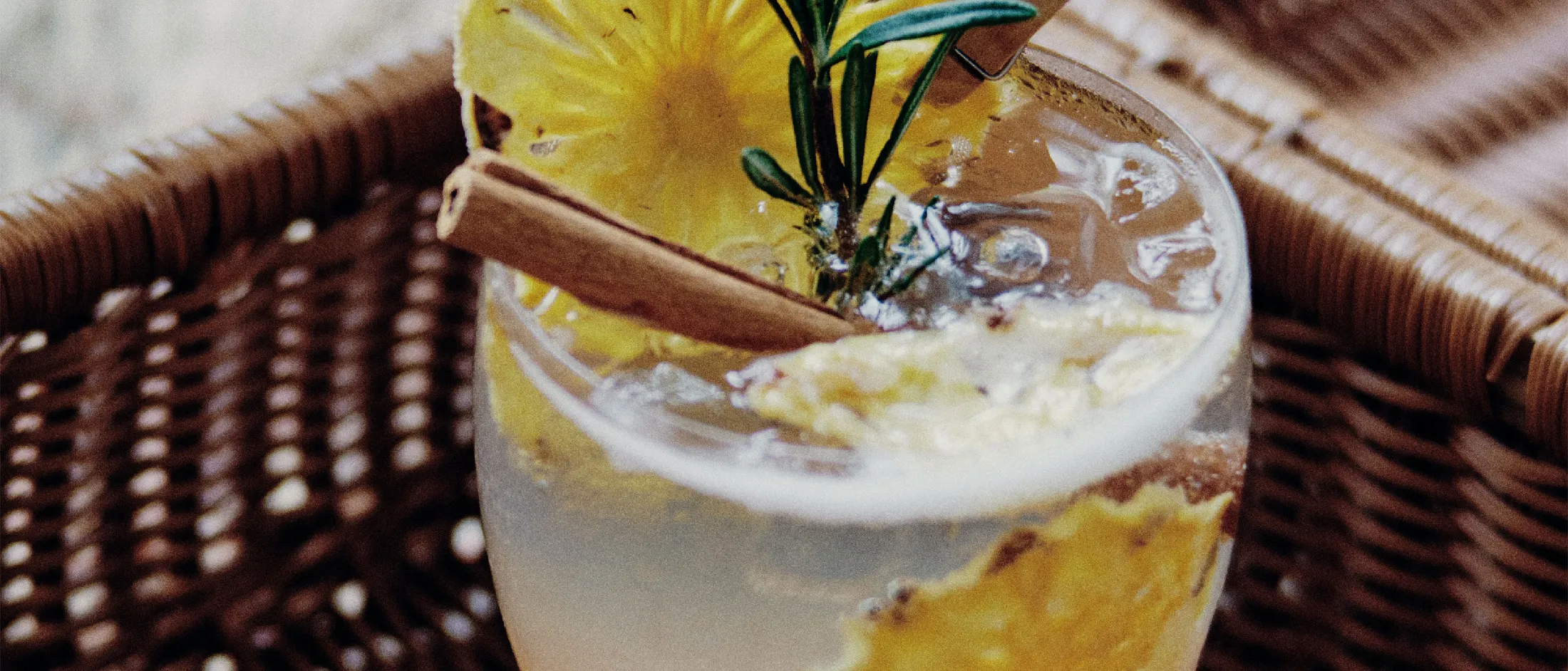 Cocktail with pineapple slices, cinnamon stick, and rosemary garnish served on a woven tray.