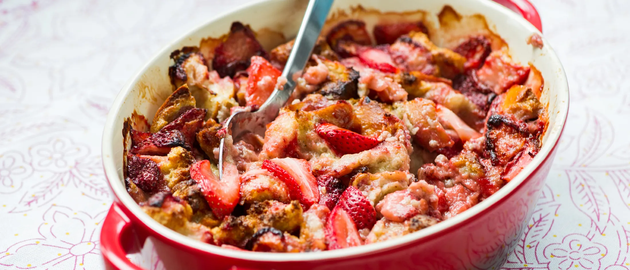 Strawberry bread pudding in a red baking dish with a spoon, on a light patterned tablecloth.