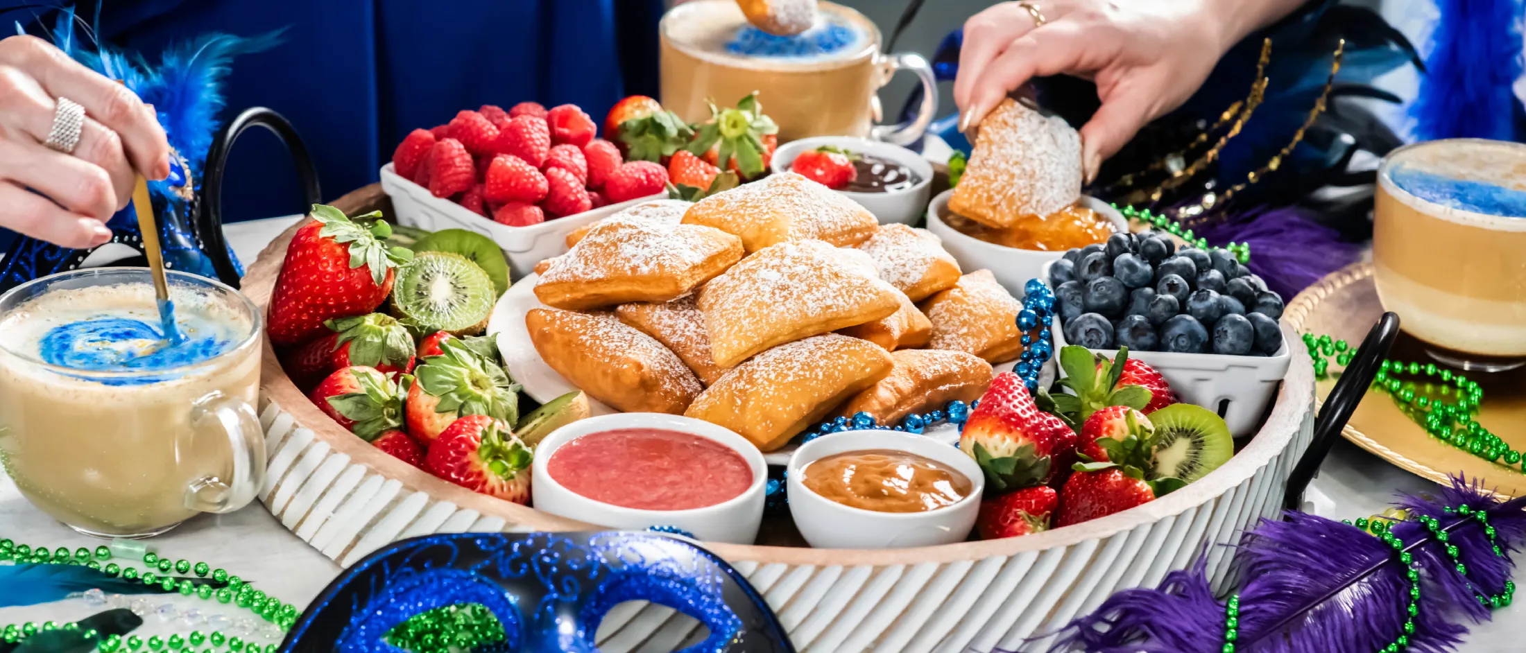 Tray of beignets, fresh berries, and dipping sauces with coffee, Mardi Gras masks, and beads on the table during a festive gathering.