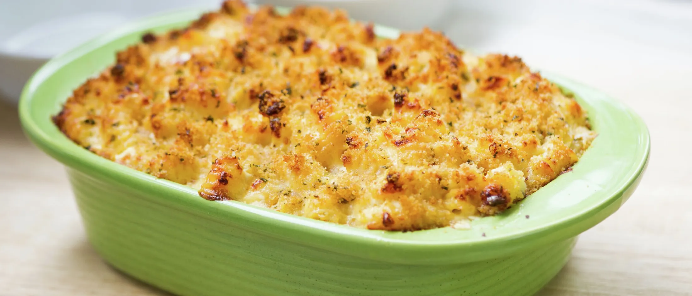 Baked macaroni and cheese with a crispy golden breadcrumb topping in a green casserole dish on a wooden surface.