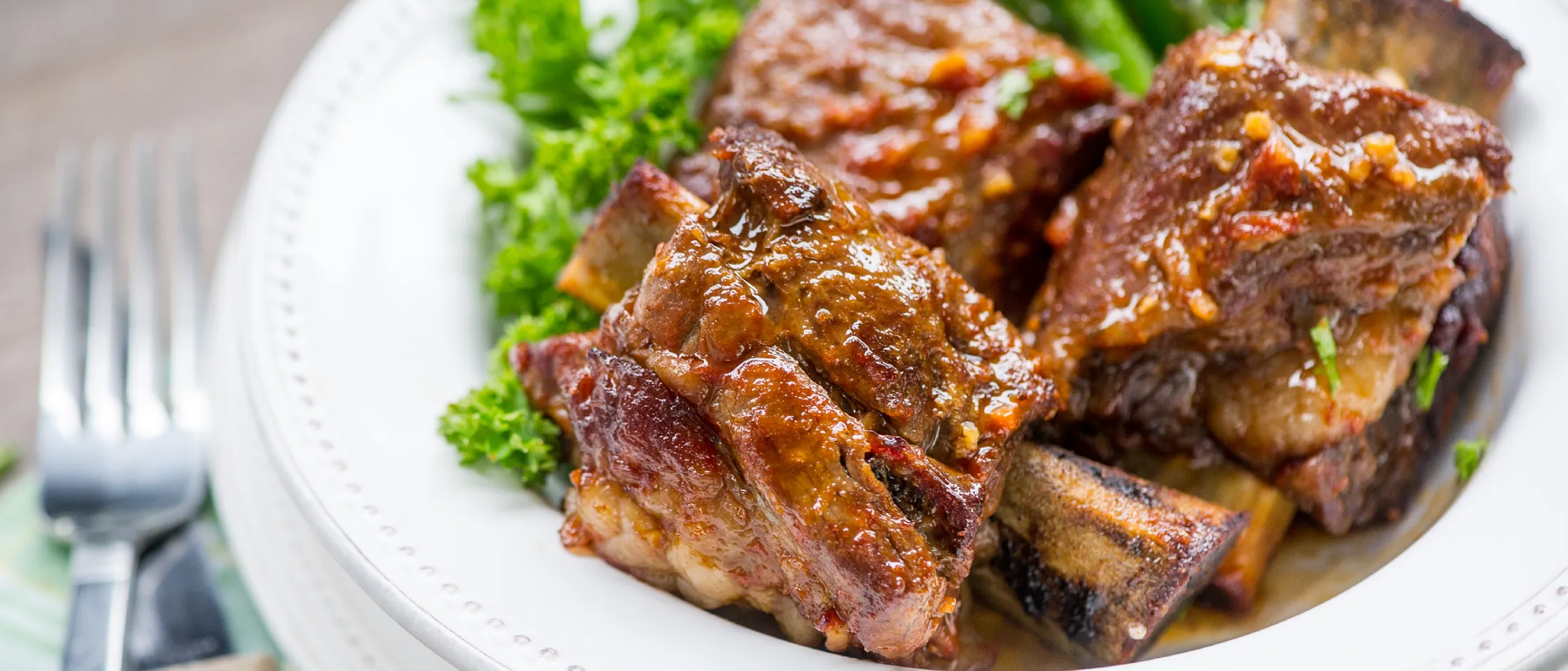 Close-up of glazed BBQ beef ribs on a white plate with green garnish and asparagus, next to a fork and knife on a napkin.