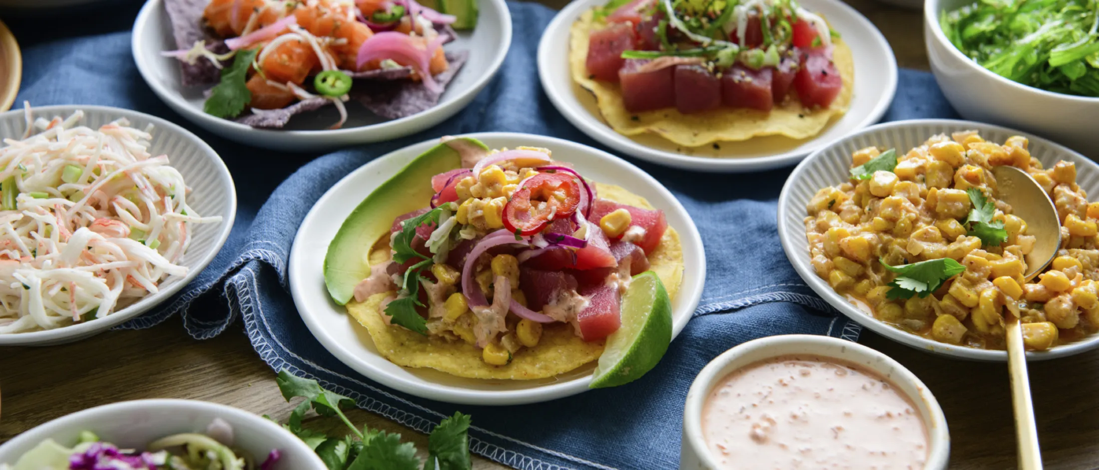 Assorted Mexican dishes on plates and bowls, including tostadas with avocado, tuna, corn salad, creamy sauce, and fresh vegetables.