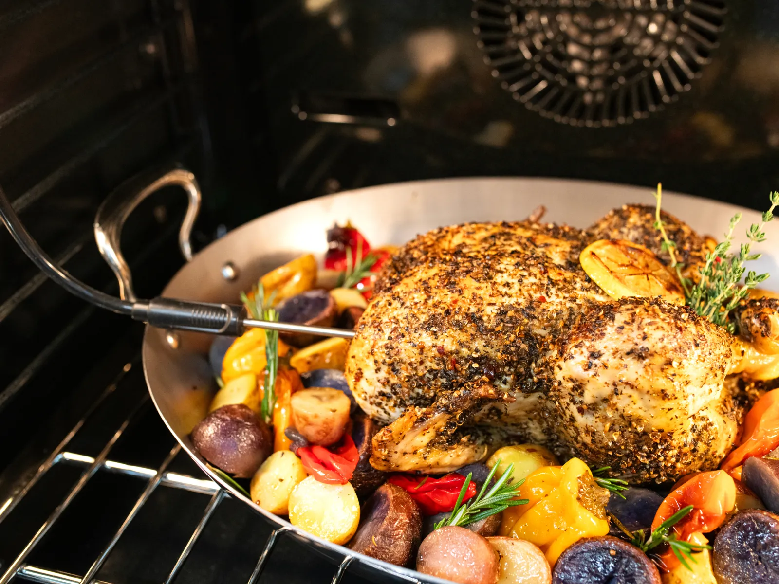 A close-up inside an oven shows a roasted chicken surrounded by colorful vegetables like potatoes, bell peppers, and herbs, all in a metal roasting pan. A meat thermometer is inserted into the chicken.