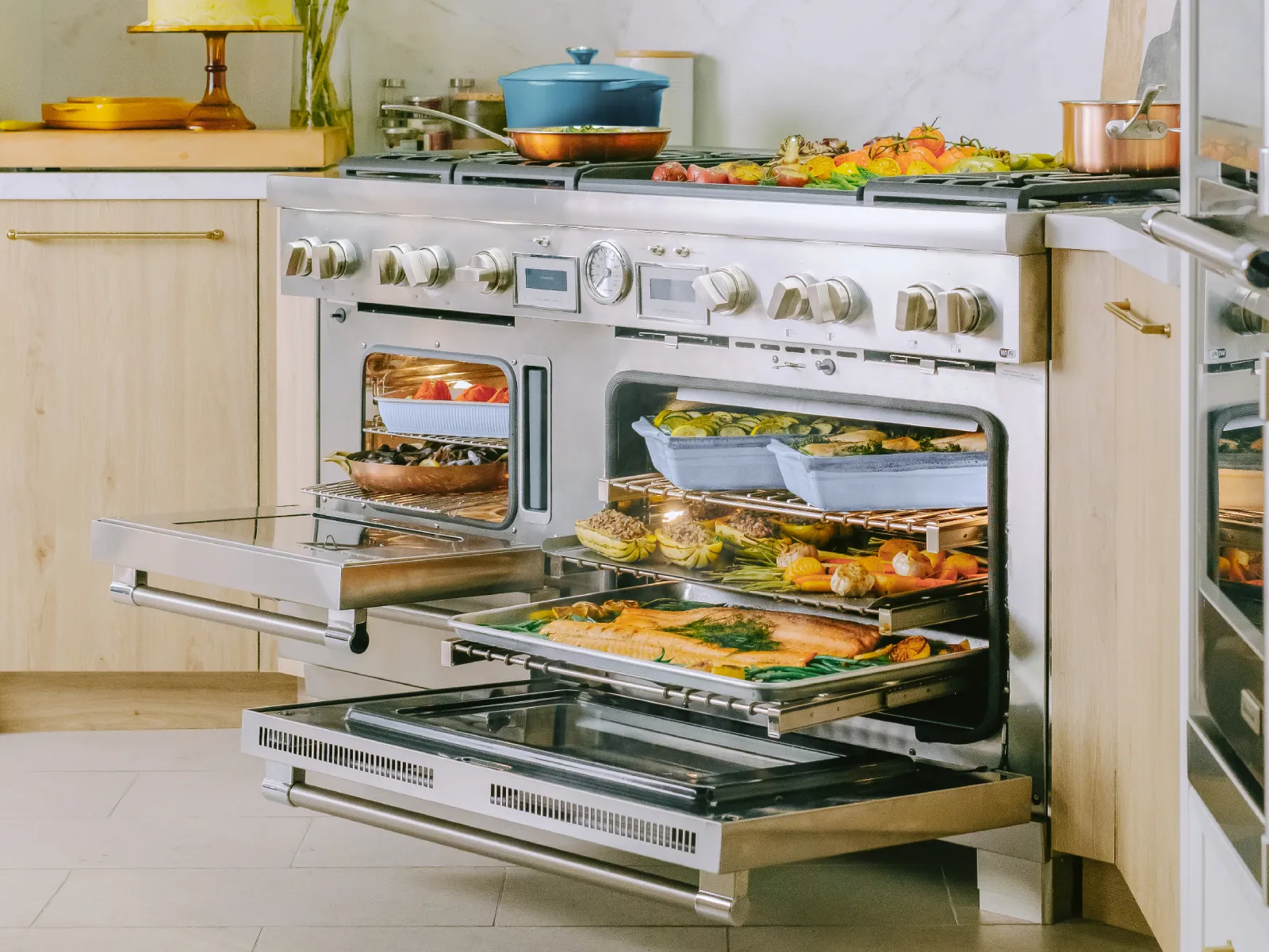 A large stainless steel range with open ovens revealing multiple dishes cooking, including fish and vegetables, with more food on the stovetop.