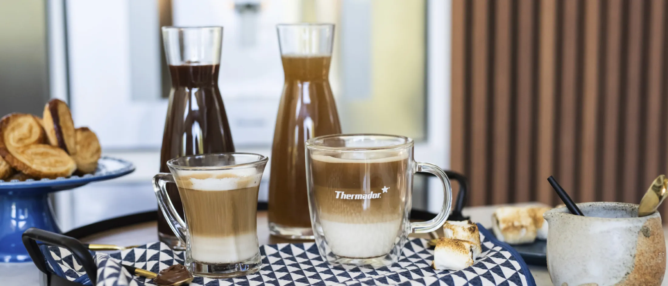 Two layered coffee mugs on a tray with carafes of syrup, marshmallows, and cookies in a cozy kitchen setting.