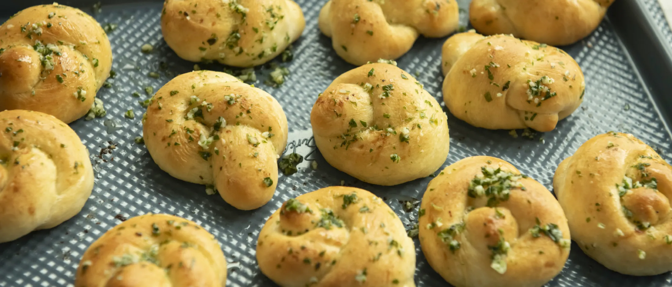Tray of freshly baked garlic herb knotted bread rolls on a textured baking sheet.