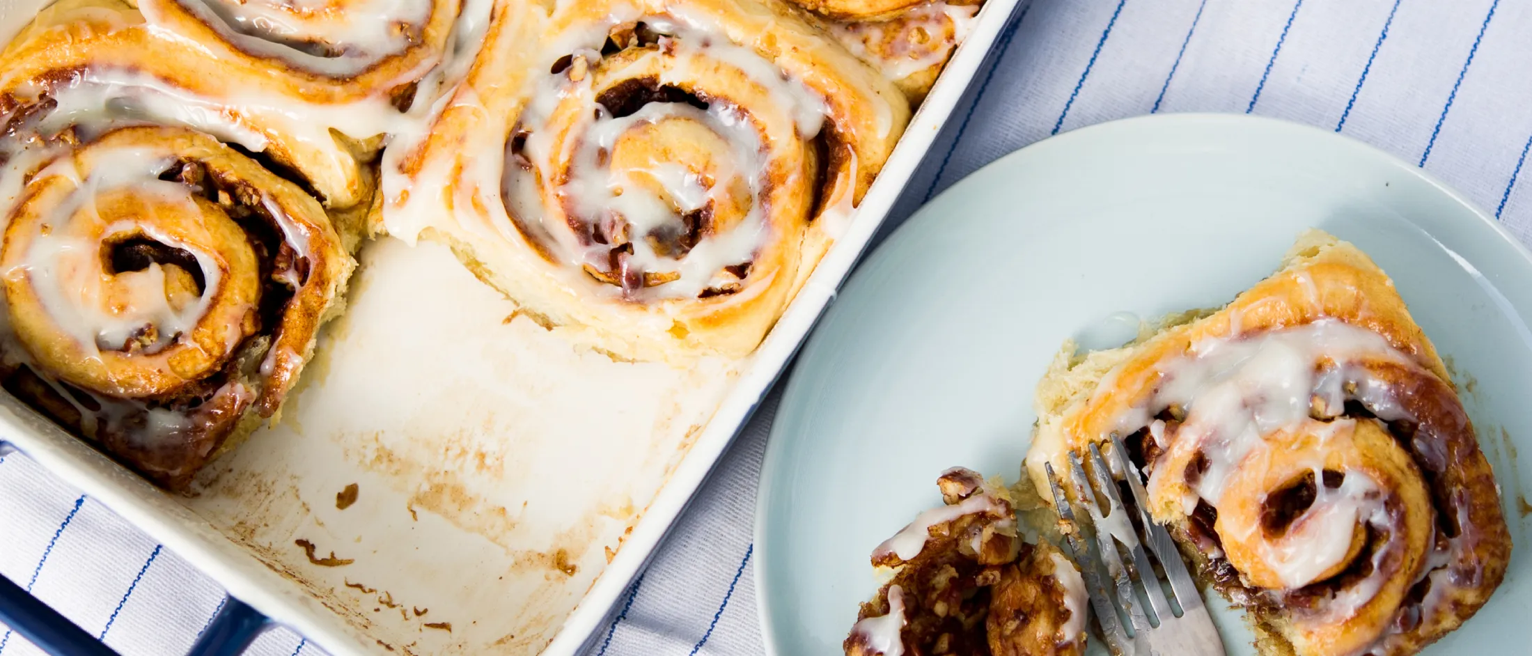 Baked cinnamon rolls with icing in a baking dish, one piece on a plate with a fork, on a striped cloth background.