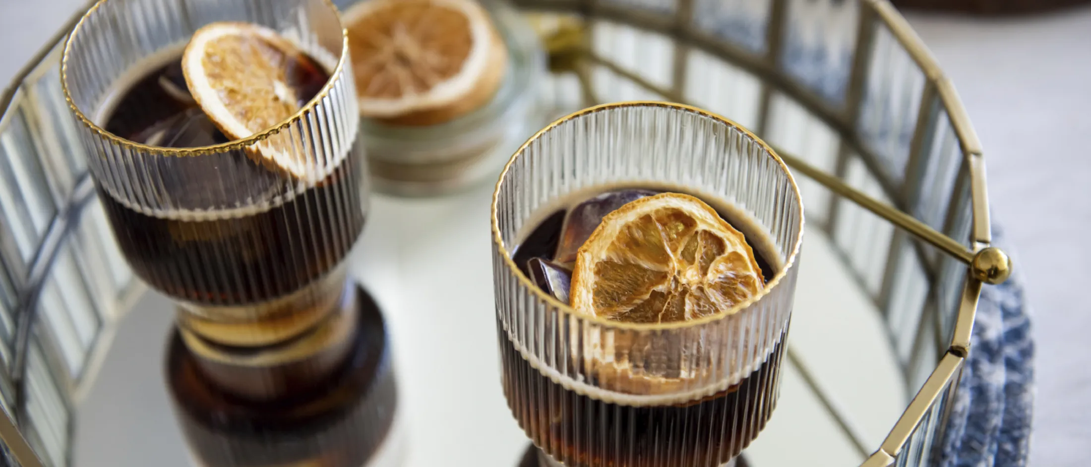 Two glasses of dark beverage garnished with dried citrus slices on a round mirrored tray with a gold rim.