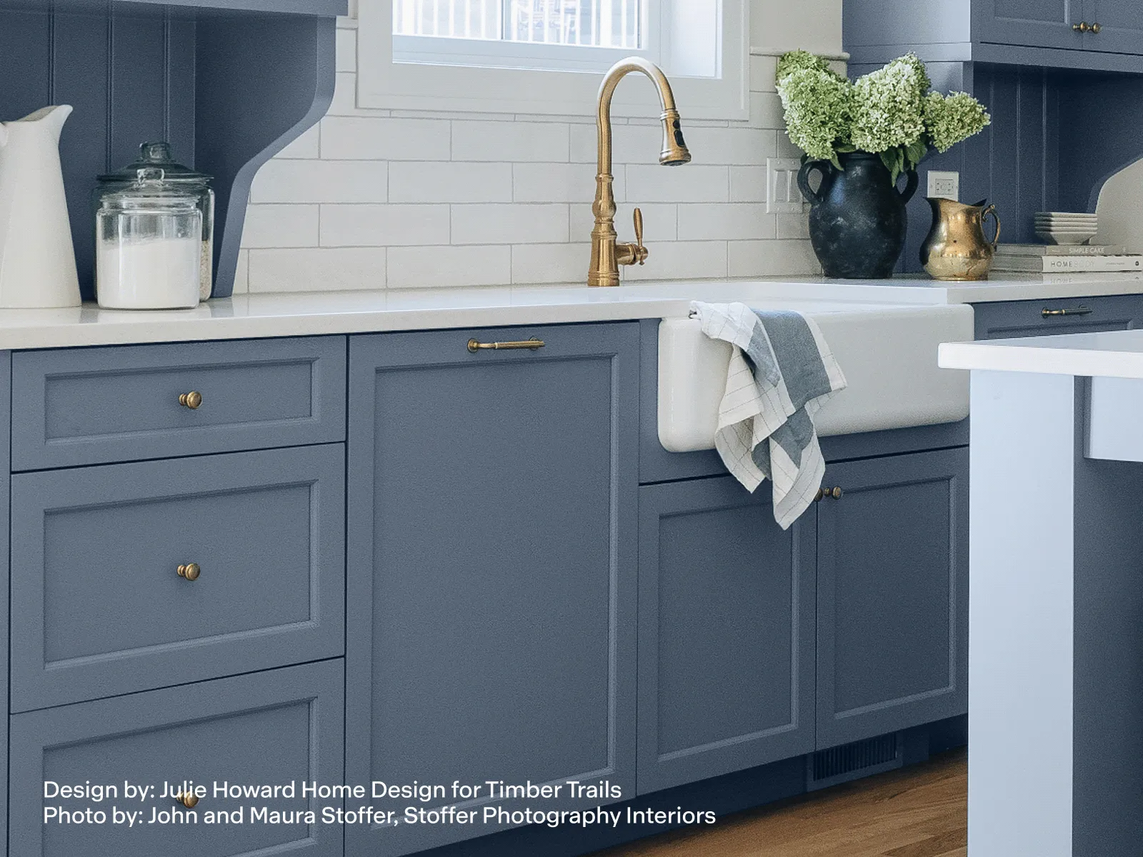 Modern kitchen with blue cabinetry, white farmhouse sink, brass faucet, white subway tile backsplash, and decorative vases with flowers.
