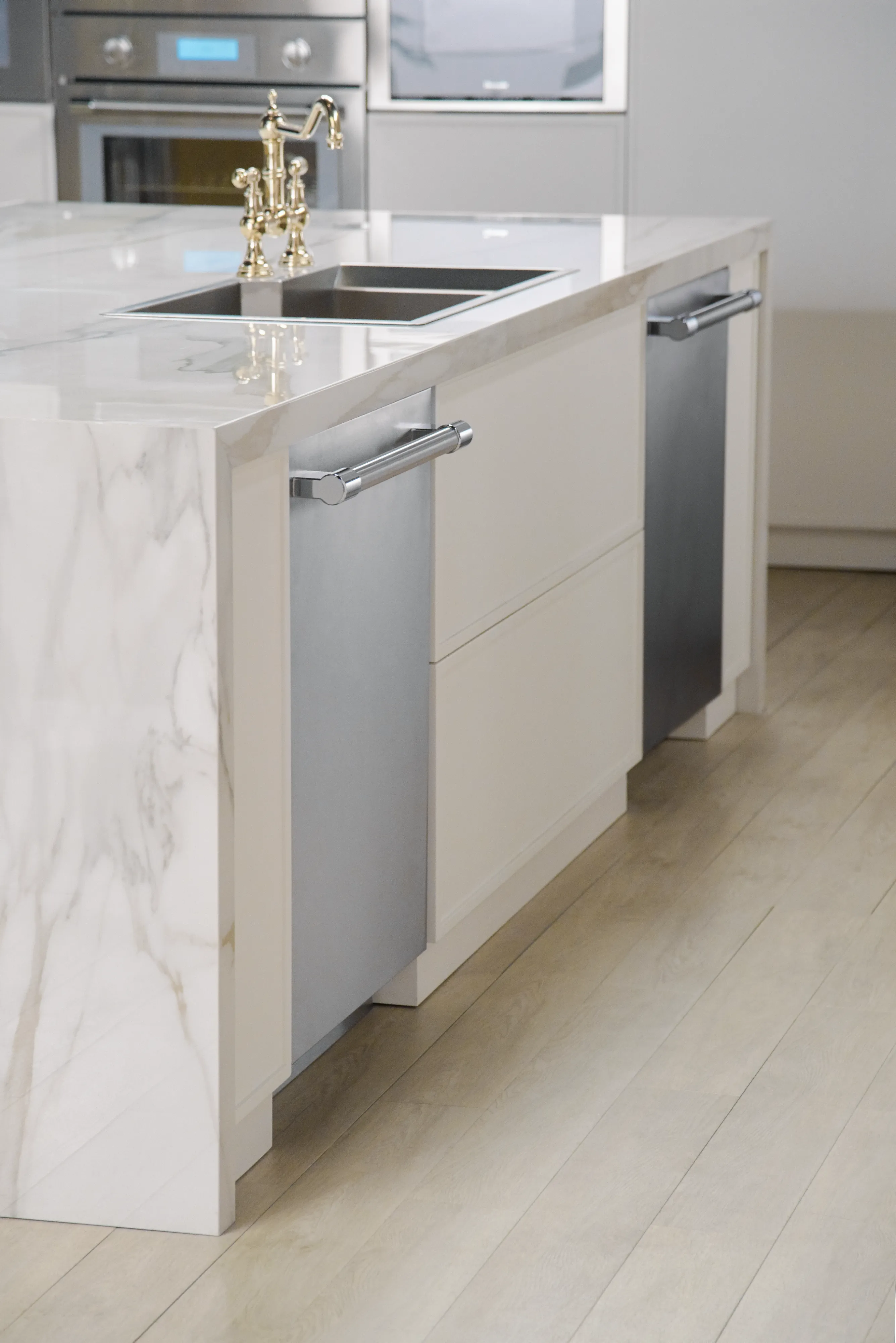 Modern kitchen island with marble countertop, dual stainless steel dishwashers, brass faucet, and built-in sink on light wood flooring.