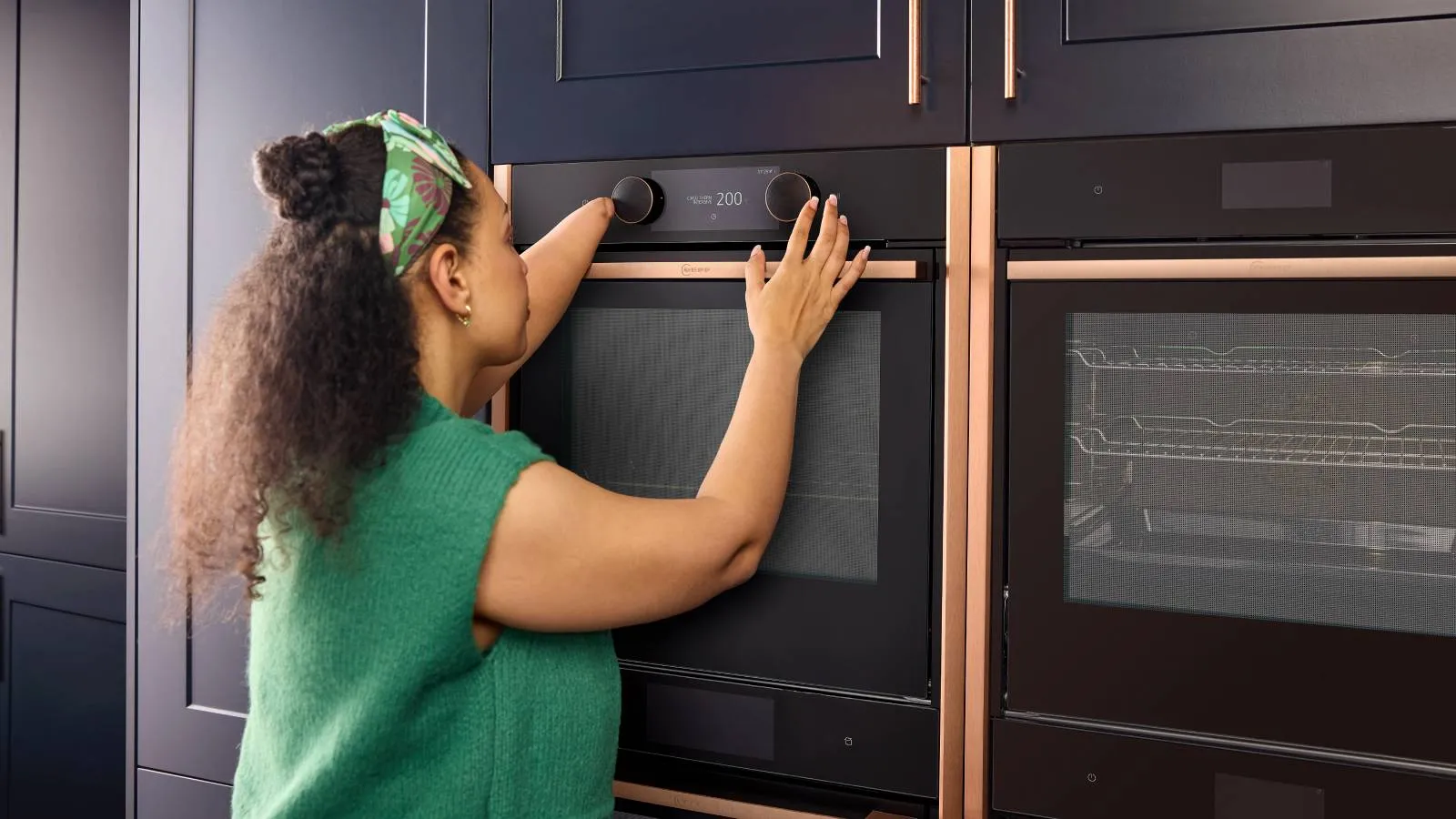 Wider shot of a woman with a limb difference affecting one forearm, from behind as she uses her arm to adjust the oven controls on the slide and hide oven.