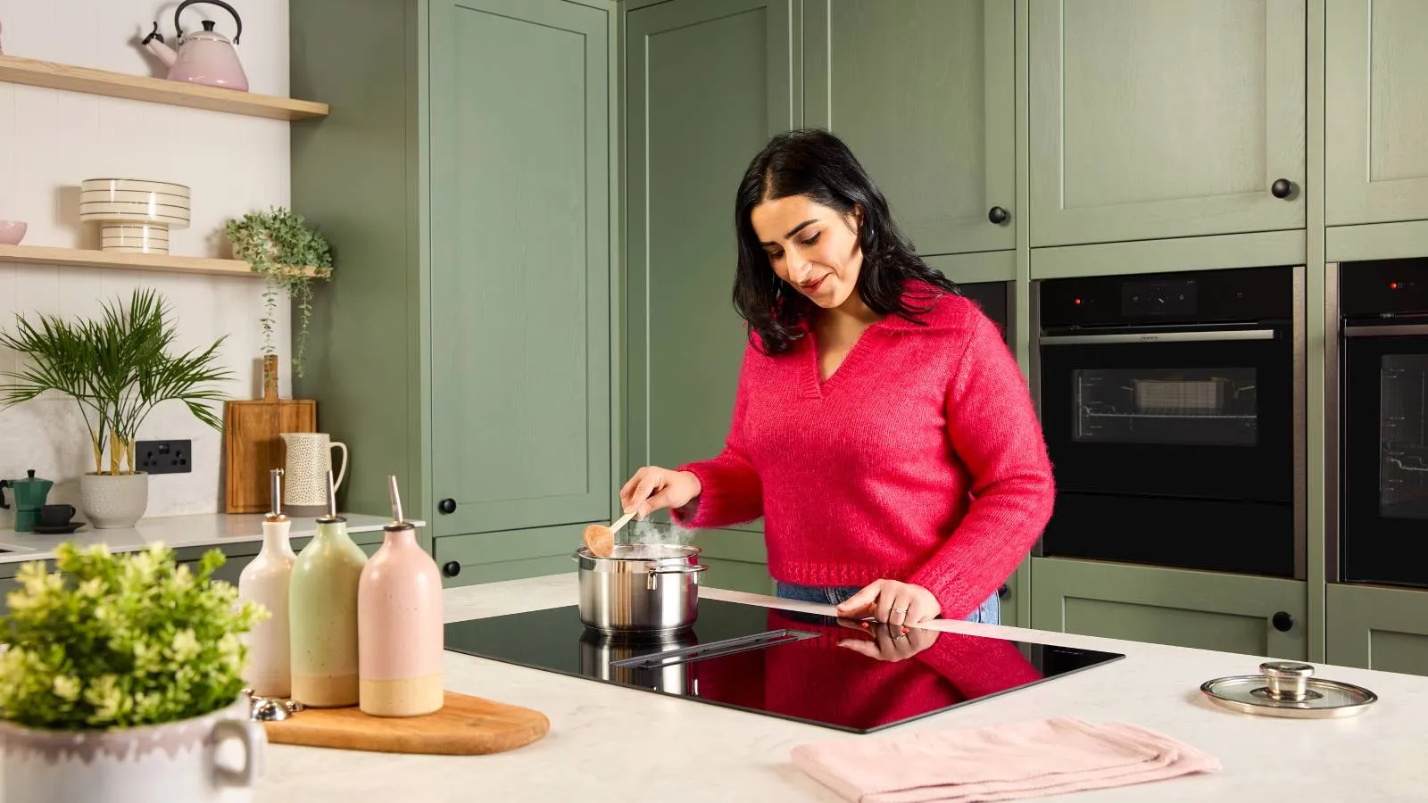 A woman who is visually impaired, wearing a bright pink sweater stirs a saucepan on a black venting induction hob, set into a white kitchen island worktop.