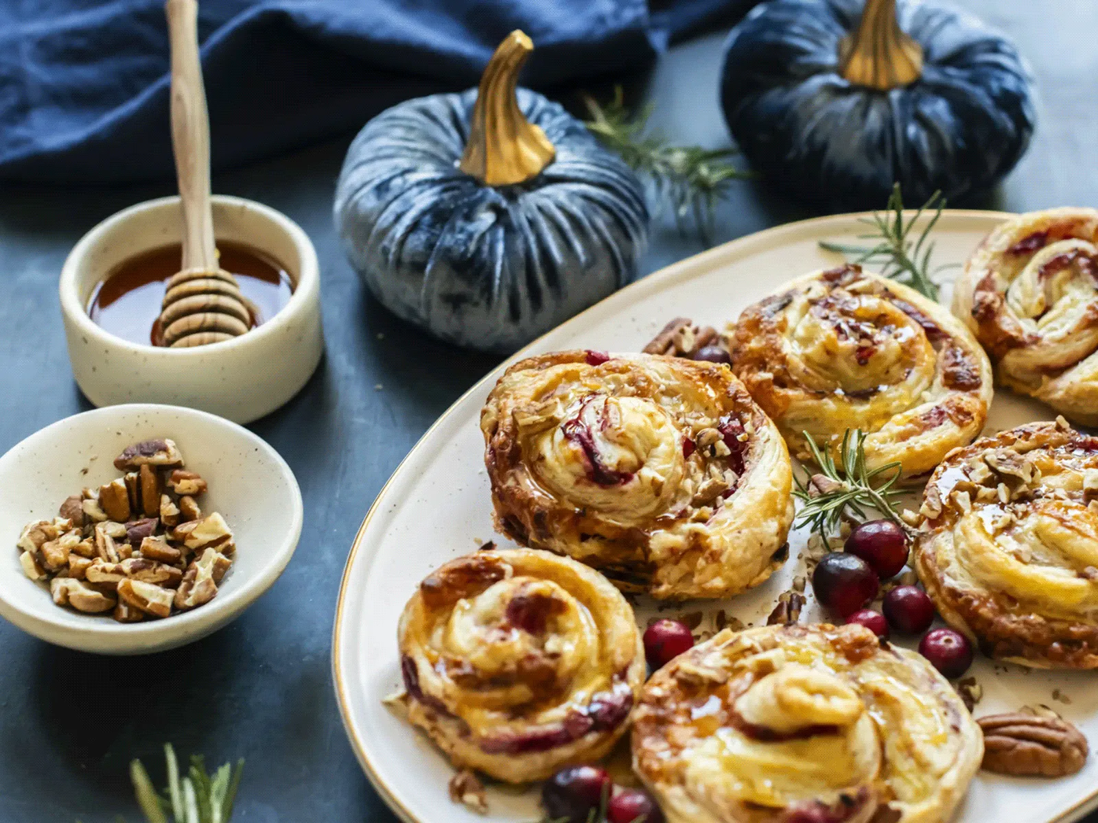 Plate of fruit-filled pastry swirls drizzled with honey and garnished with cranberries and rosemary, with bowls of honey and nuts and decorative pumpkins nearby.