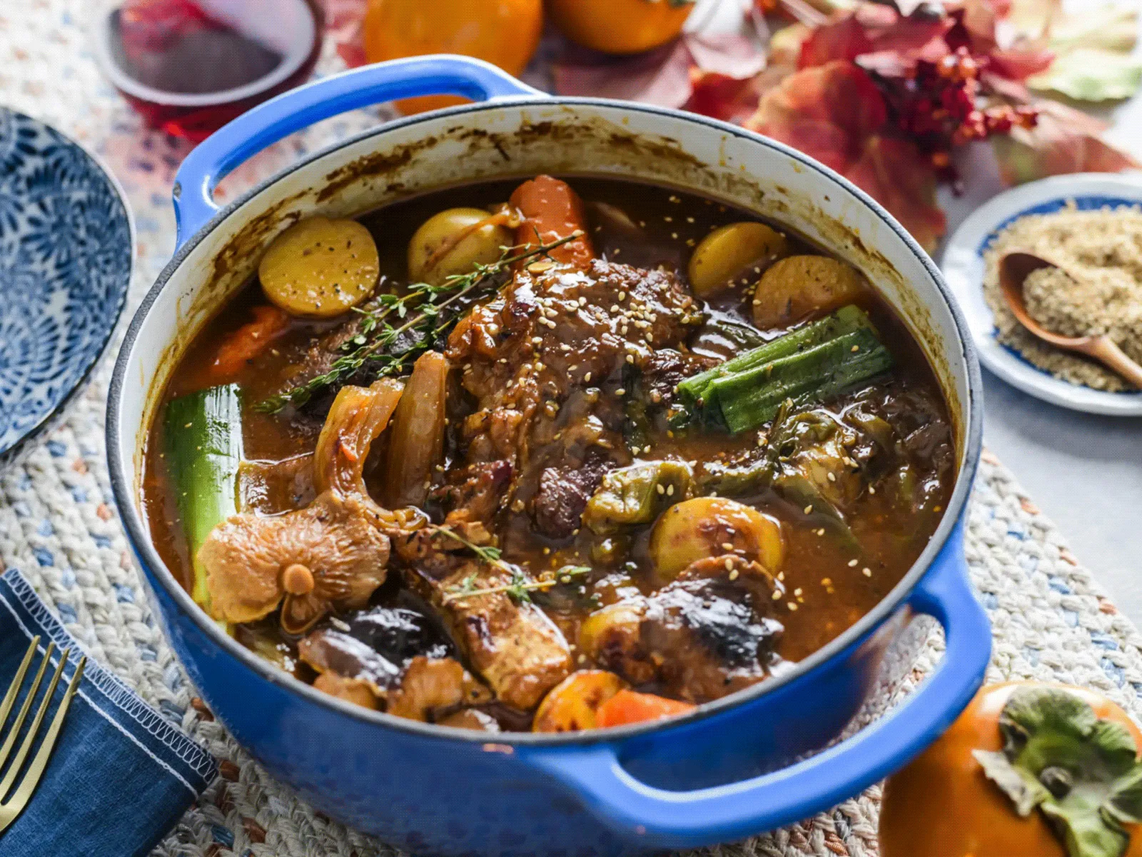 Hearty stew with chicken, okra, and vegetables in a blue pot, surrounded by blue plates and garnishes on a patterned tablecloth.