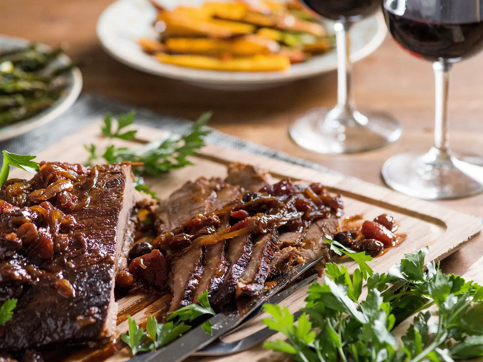 Sliced grilled steak topped with caramelized onions and parsley on a wooden board, with roasted vegetables and two glasses of red wine in the background.