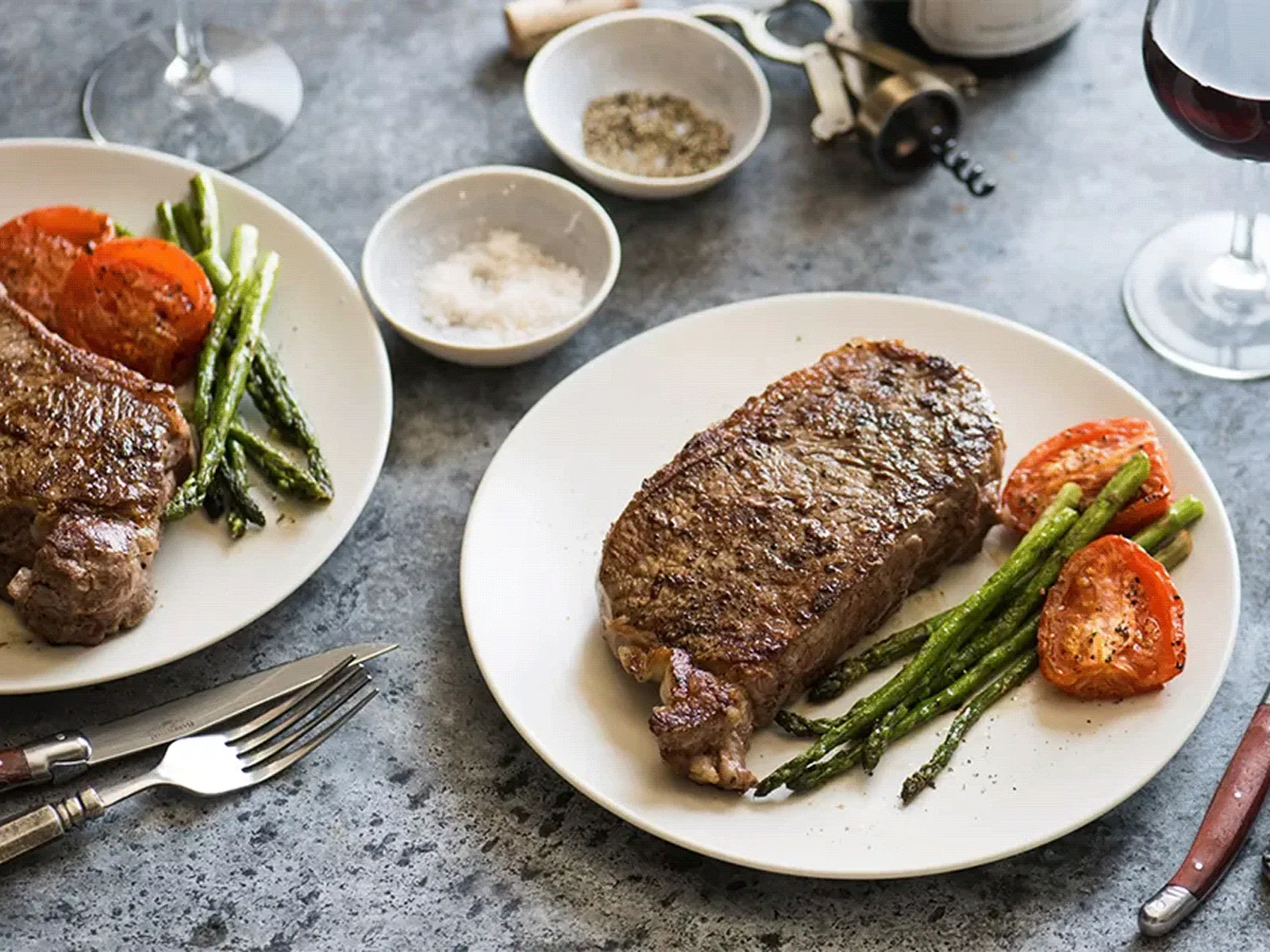 Two plates of seared steak with asparagus and roasted tomatoes, with salt and pepper bowls and glasses of red wine.