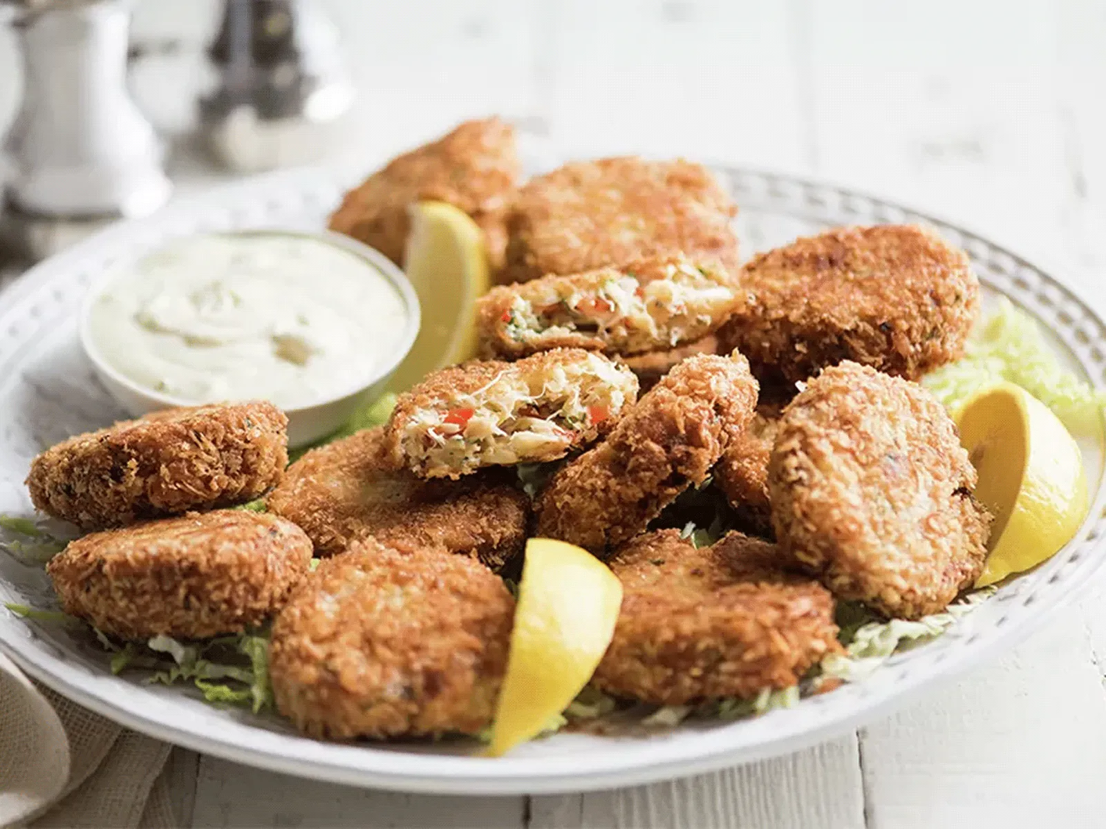 Plate of golden-brown crab cakes on lettuce with lemon wedges and a small bowl of creamy dipping sauce.