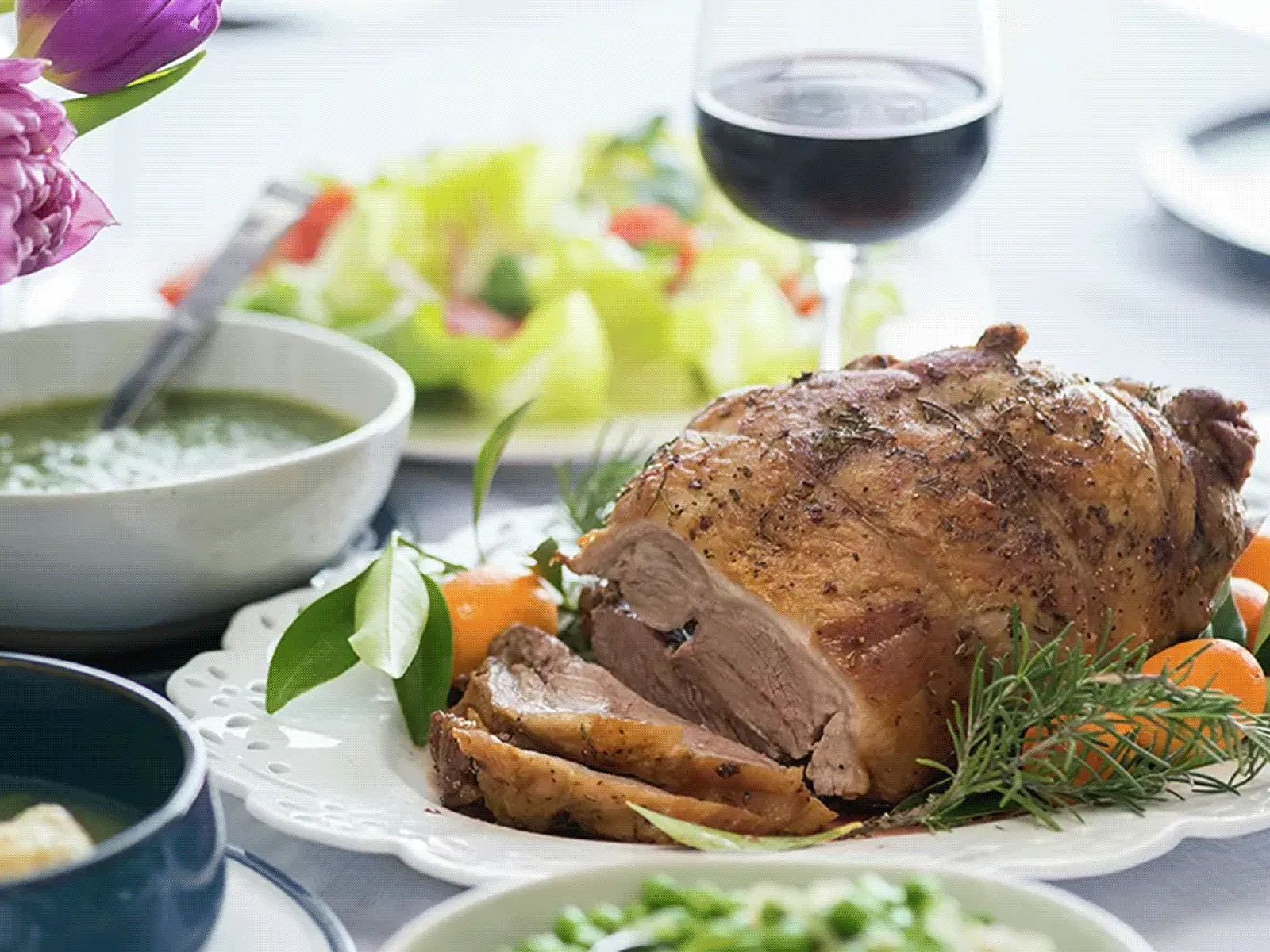 Roasted leg of lamb sliced on a white platter with herbs and carrots, surrounded by bowls of salad, peas, a green sauce, and a glass of red wine.