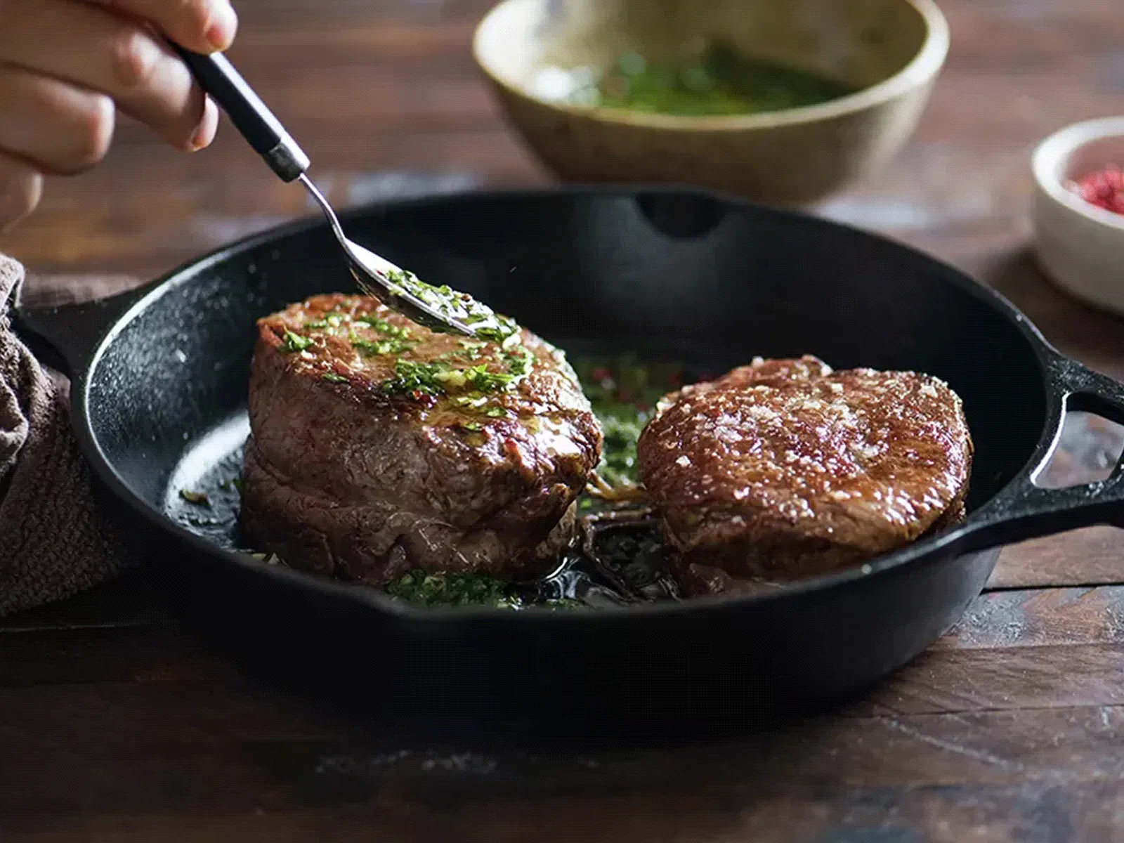 Two seared steak medallions in a cast-iron skillet, sprinkled with chopped herbs as a hand presses one with a fork, with small bowls of sauce and seasoning in the background.