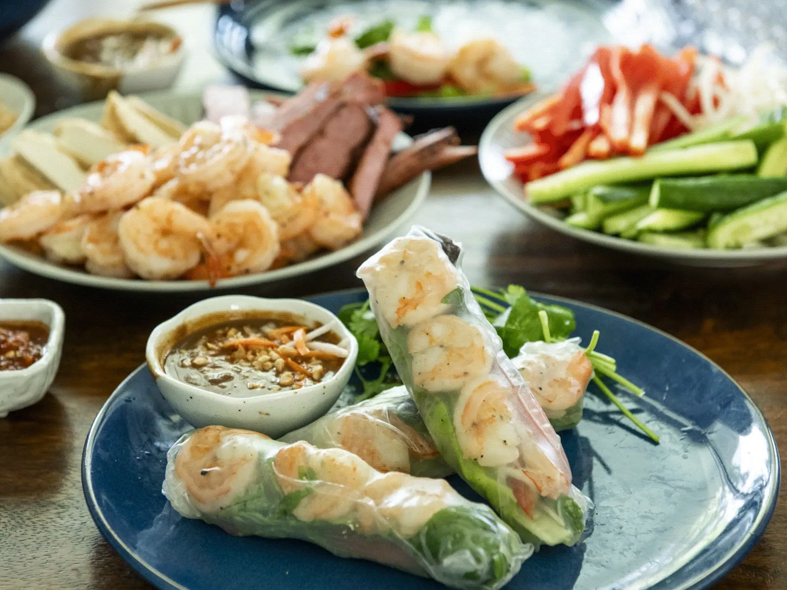 Fresh shrimp spring rolls on a blue plate with a small bowl of dipping sauce, surrounded by plates of shrimp, sliced cucumber, and other vegetables.