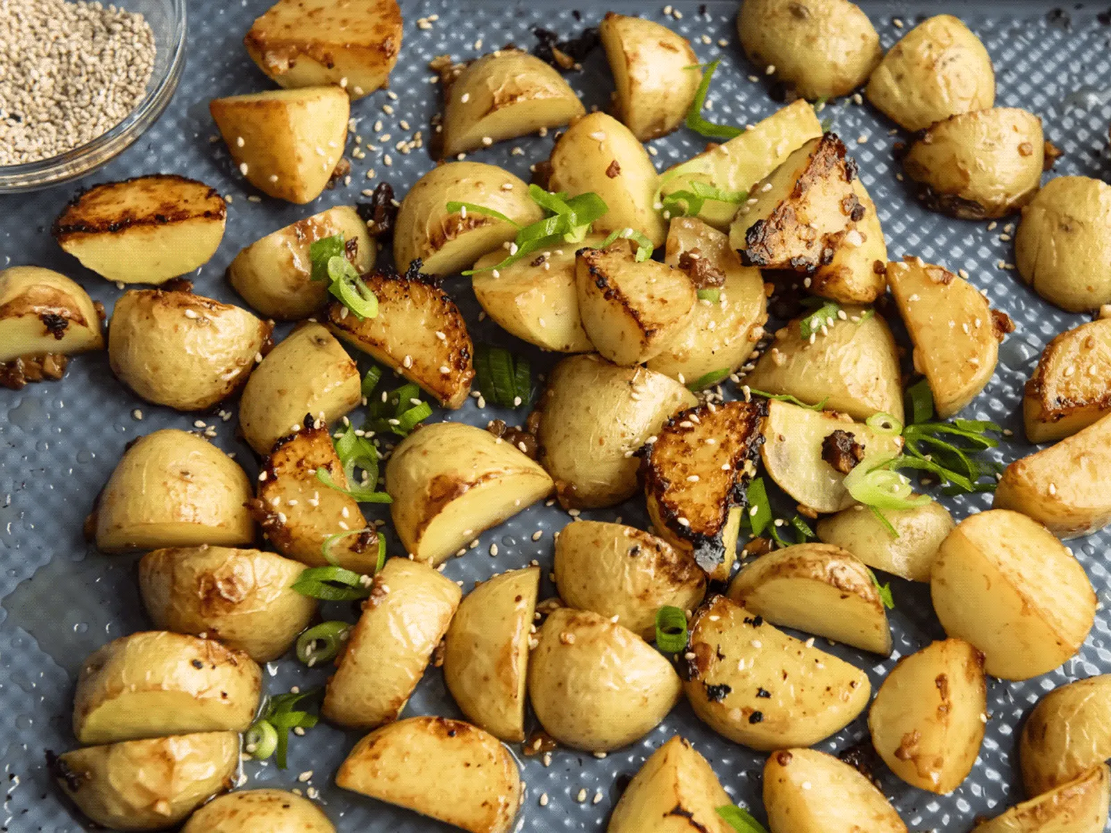 Roasted baby potato wedges seasoned with sesame seeds and sliced green onions on a baking sheet.