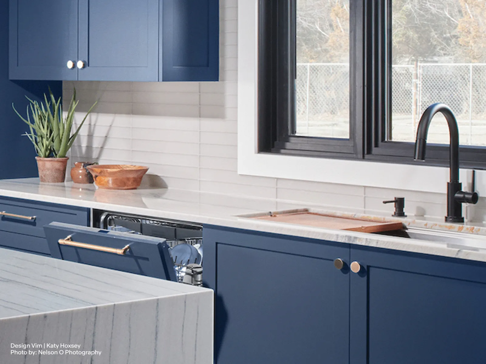 Modern kitchen featuring blue cabinets, white countertops, a black faucet, and a window overlooking an outdoor area.