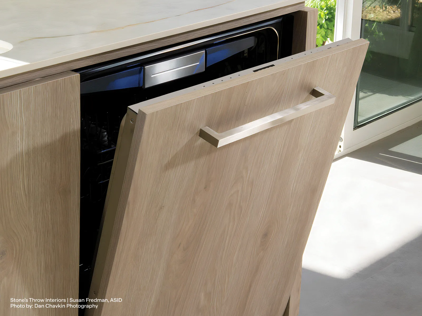 Partially open integrated dishwasher with a light wood cabinet front in a sunlit modern kitchen.