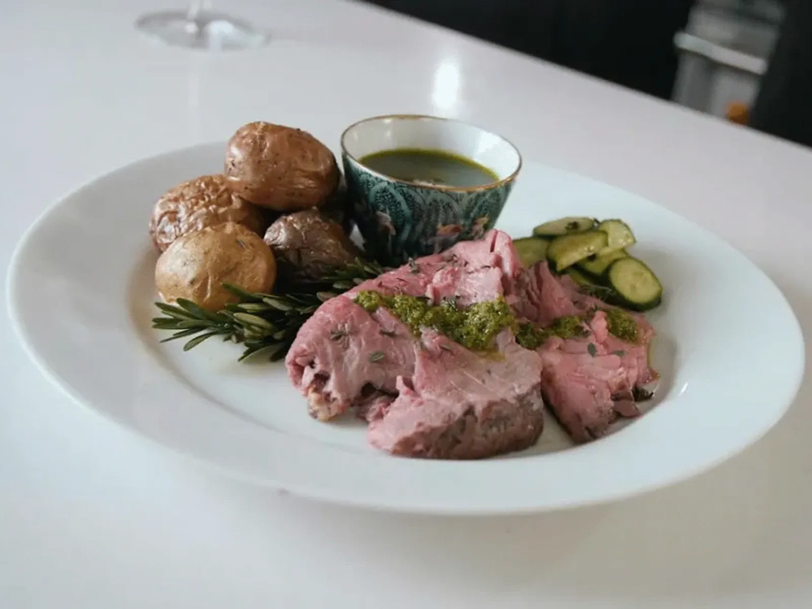 Plate with sliced roast beef, roasted baby potatoes, zucchini slices, and a small cup of green herb sauce, garnished with rosemary.