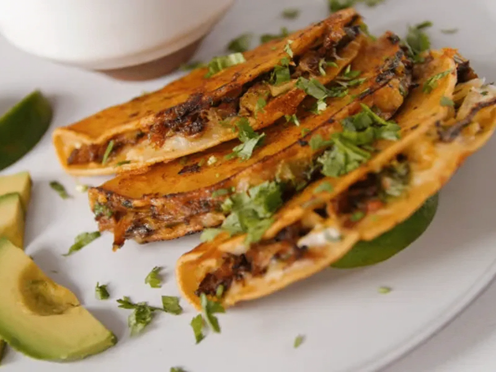 Three small corn tacos with savory filling and chopped cilantro, served with lime wedge and avocado slices on a white plate.