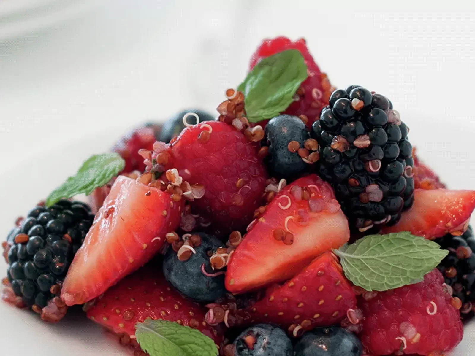 Mixed berry salad with strawberries, raspberries, blackberries, blueberries, quinoa, and fresh mint leaves on a white plate.