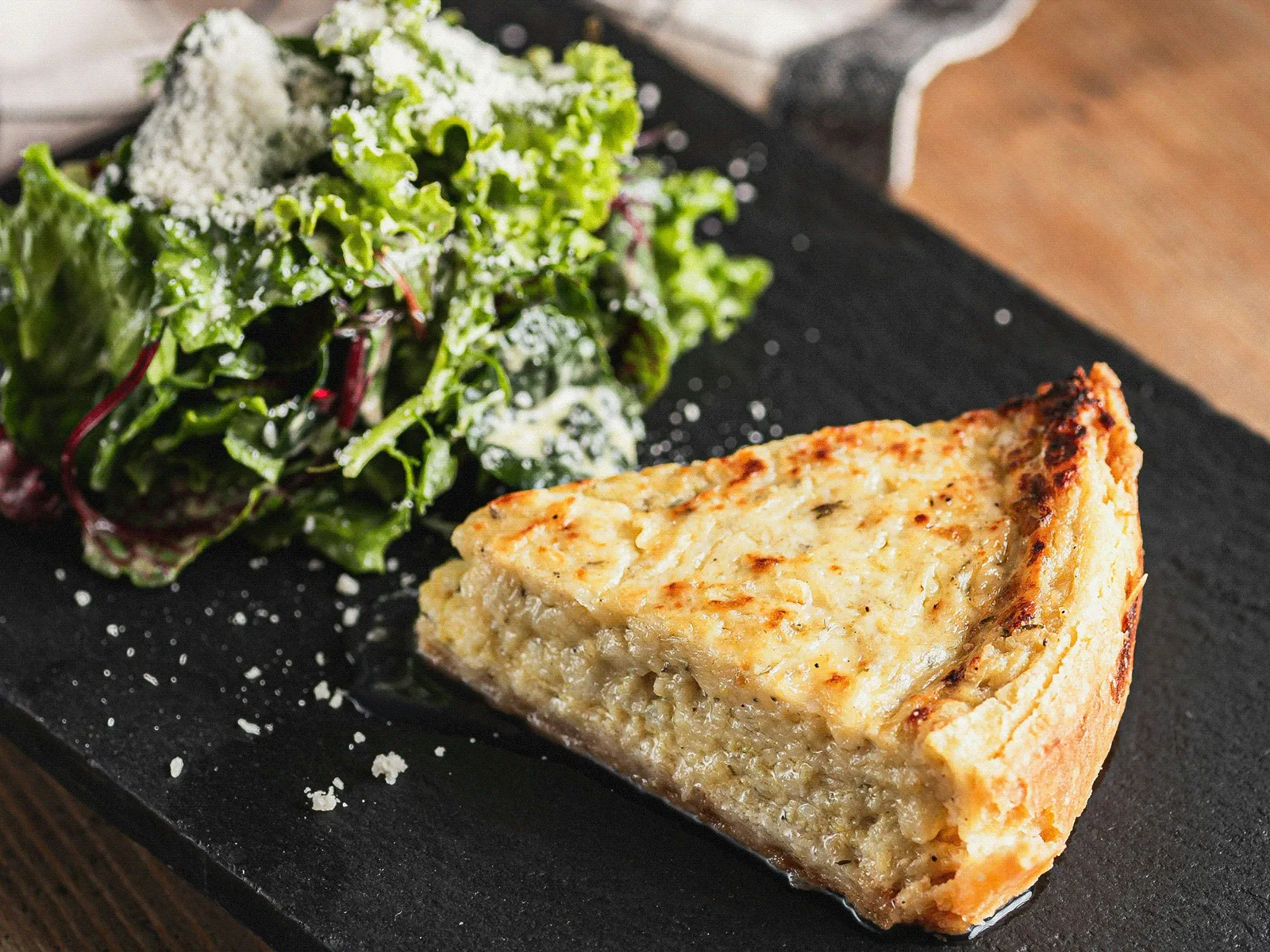 Slice of golden quiche on a black slate board with a side of leafy green salad dusted with grated cheese and a checked napkin in the background.