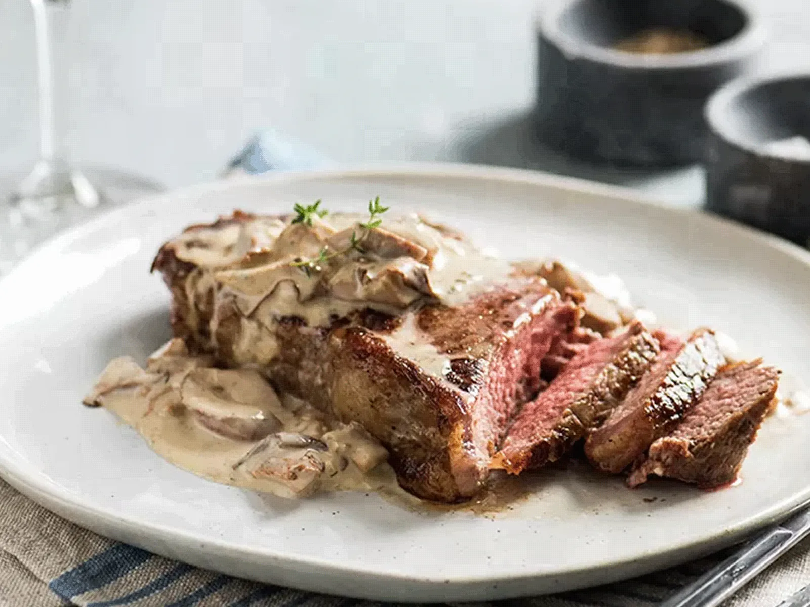 Sliced steak with creamy mushroom sauce on a white plate, with a glass of red wine in the background.