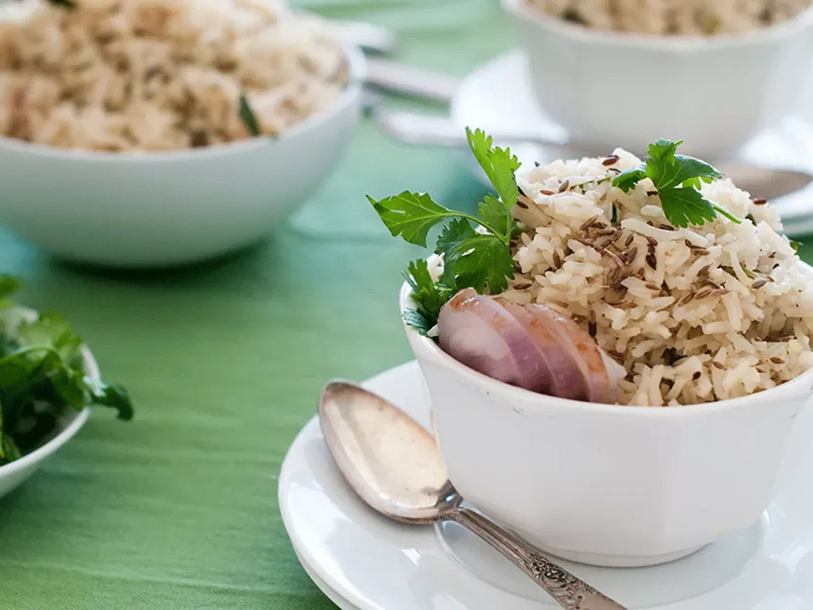 Bowl of seasoned rice garnished with parsley and a shallot slice, with extra bowls and a spoon on a green tablecloth.