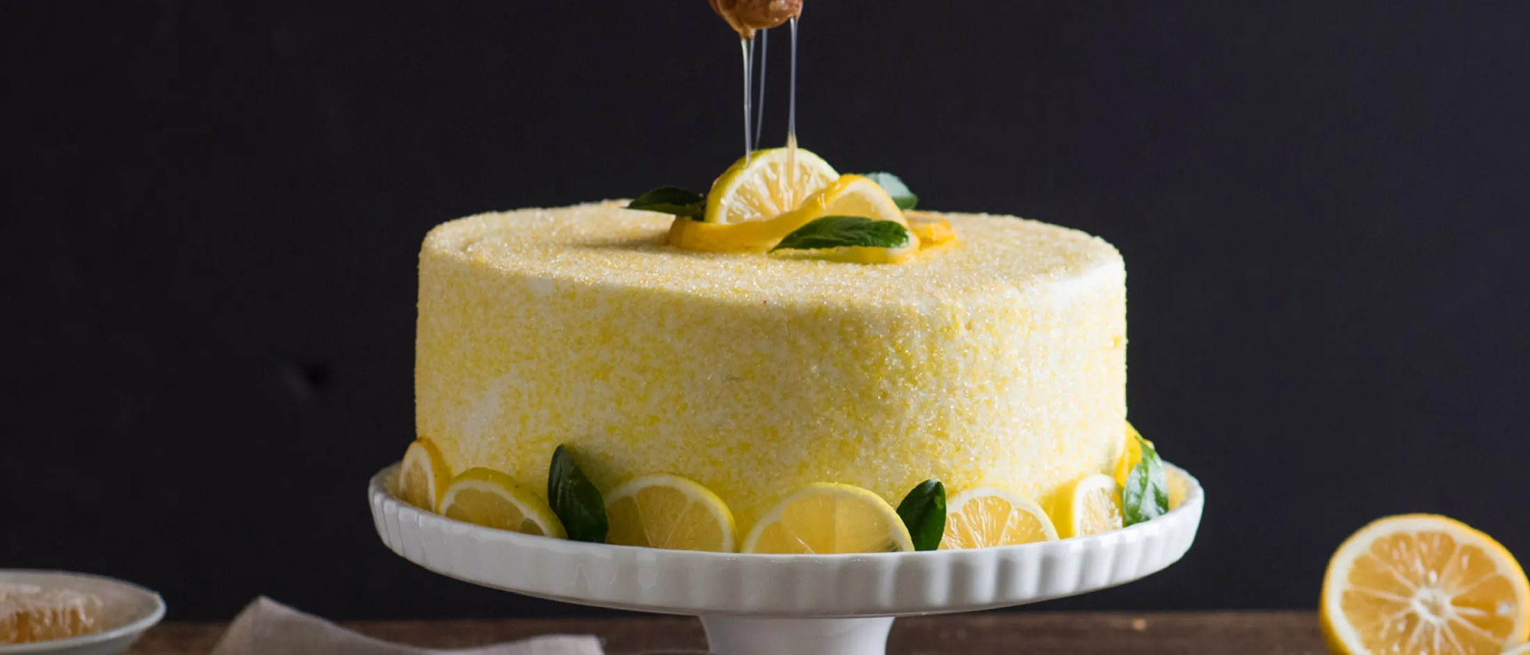 Lemon cake with fresh lemon slices and mint leaves on a white cake stand against a dark background.