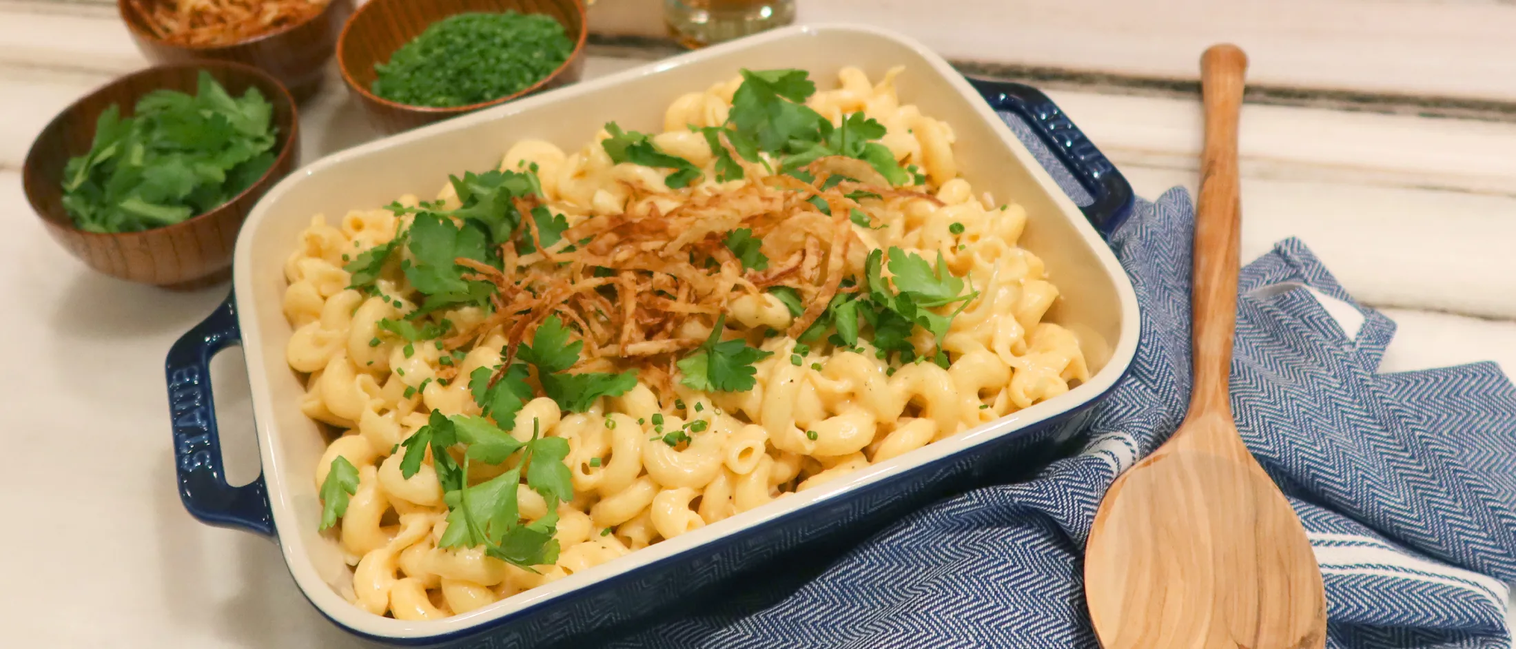 Baked macaroni and cheese garnished with fried onions and fresh herbs, served in a blue-handled dish with wooden bowls of toppings and glasses in the background