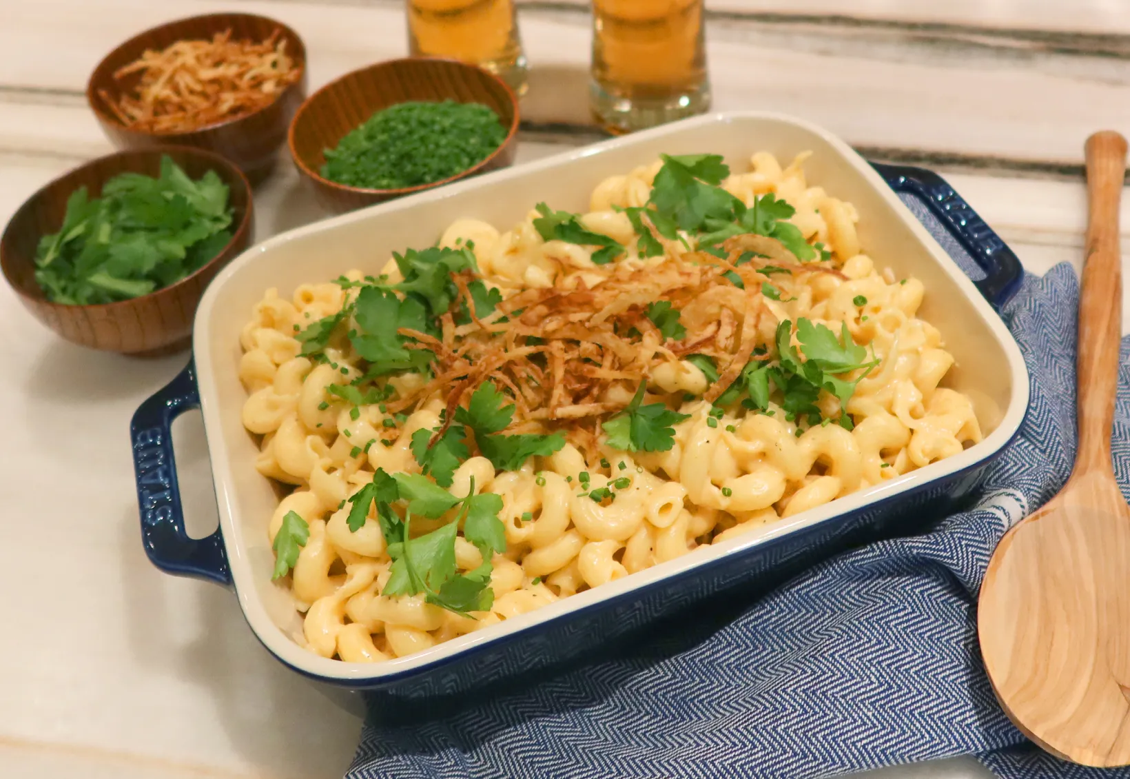 Baked macaroni and cheese garnished with fried onions and fresh herbs, served in a blue-handled dish with wooden bowls of toppings and glasses in the background